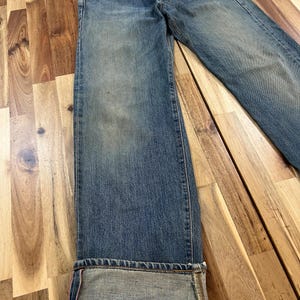 May include: Faded blue denim jeans with a button fly and a classic five-pocket design. The jeans have a relaxed fit and are cuffed at the hem, revealing a contrasting fabric. The jeans are displayed on a wooden surface.