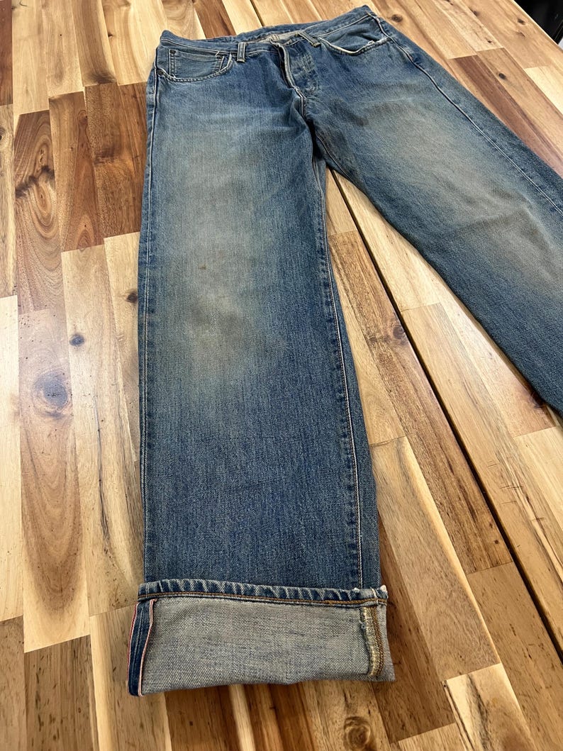 May include: Faded blue denim jeans with a button fly and a classic five-pocket design. The jeans have a relaxed fit and are cuffed at the hem, revealing a contrasting fabric. The jeans are displayed on a wooden surface.