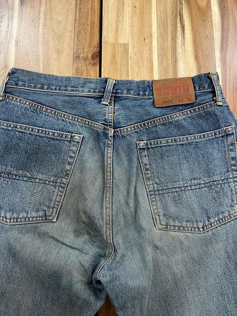 May include: Close-up of the back of blue denim jeans. The jeans have two back pockets and a brown leather patch with the brand name "EDWIN". The jeans are labeled "505X 33 34".