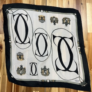 May include: A square silk scarf with a white background, featuring black interlocking "C" shapes and ornate crests. The scarf has a black border and a tassel at one corner. The text "CARTIER PARIS" is visible.