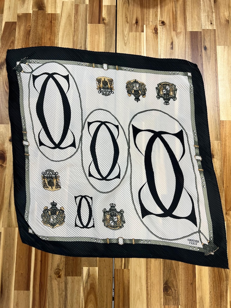 May include: A square silk scarf with a white background, featuring black interlocking "C" shapes and ornate crests. The scarf has a black border and a tassel at one corner. The text "CARTIER PARIS" is visible.