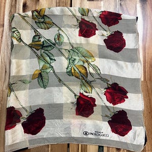 May include: A square scarf with a checkered pattern in shades of gray and white. The scarf is adorned with a repeating design of red roses with green leaves and stems. The brand name "PAOLOGUCCI" is visible.