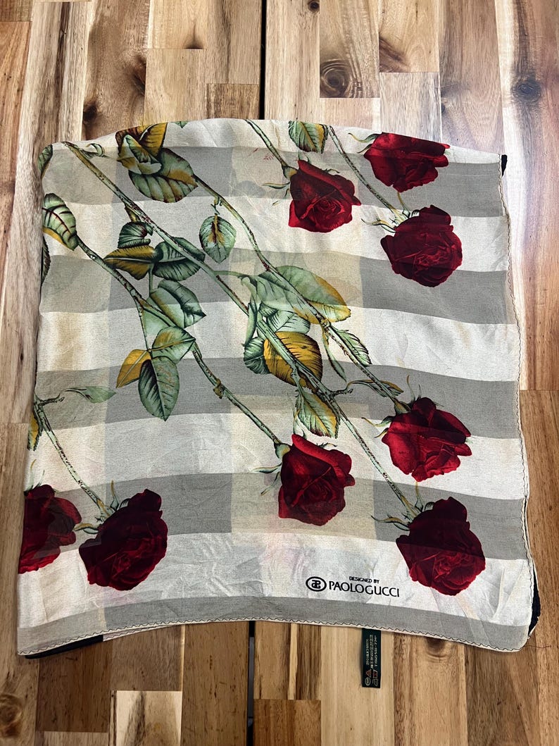 May include: A square scarf with a checkered pattern in shades of gray and white. The scarf is adorned with a repeating design of red roses with green leaves and stems. The brand name "PAOLOGUCCI" is visible.