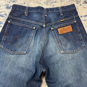 Może przedstawiać: Zbliżenie na tył niebieskich dżins&oacute;w Wrangler. Dżinsy mają dwie tylne kieszenie z charakterystycznym wzorem szw&oacute;w, brązową sk&oacute;rzaną naszywkę z logo Wrangler i klasyczny kr&oacute;j.