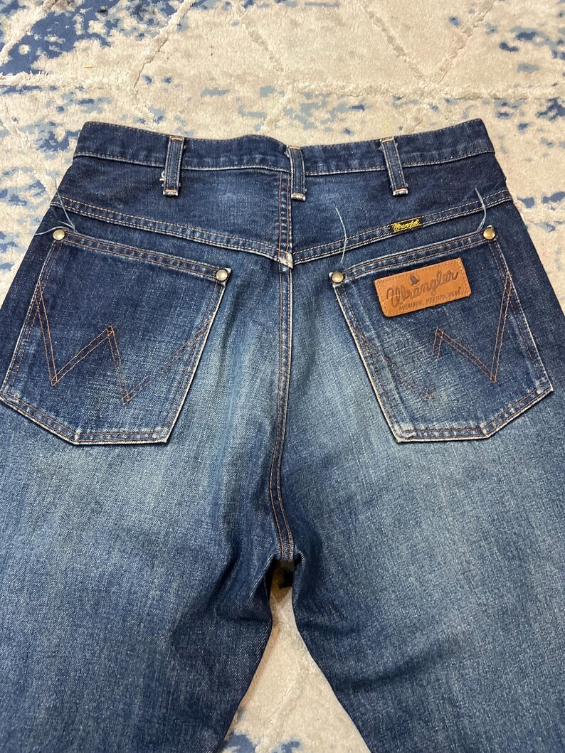 Może przedstawiać: Zbliżenie na tył niebieskich dżins&oacute;w Wrangler. Dżinsy mają dwie tylne kieszenie z charakterystycznym wzorem szw&oacute;w, brązową sk&oacute;rzaną naszywkę z logo Wrangler i klasyczny kr&oacute;j.