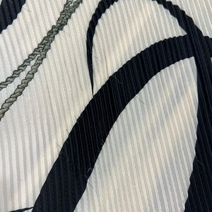 May include: Close-up of a fabric with a pleated design. The fabric features a pattern of vertical white stripes, with bold black curved shapes and a gray braided detail. The texture is clearly visible, highlighting the pleats.