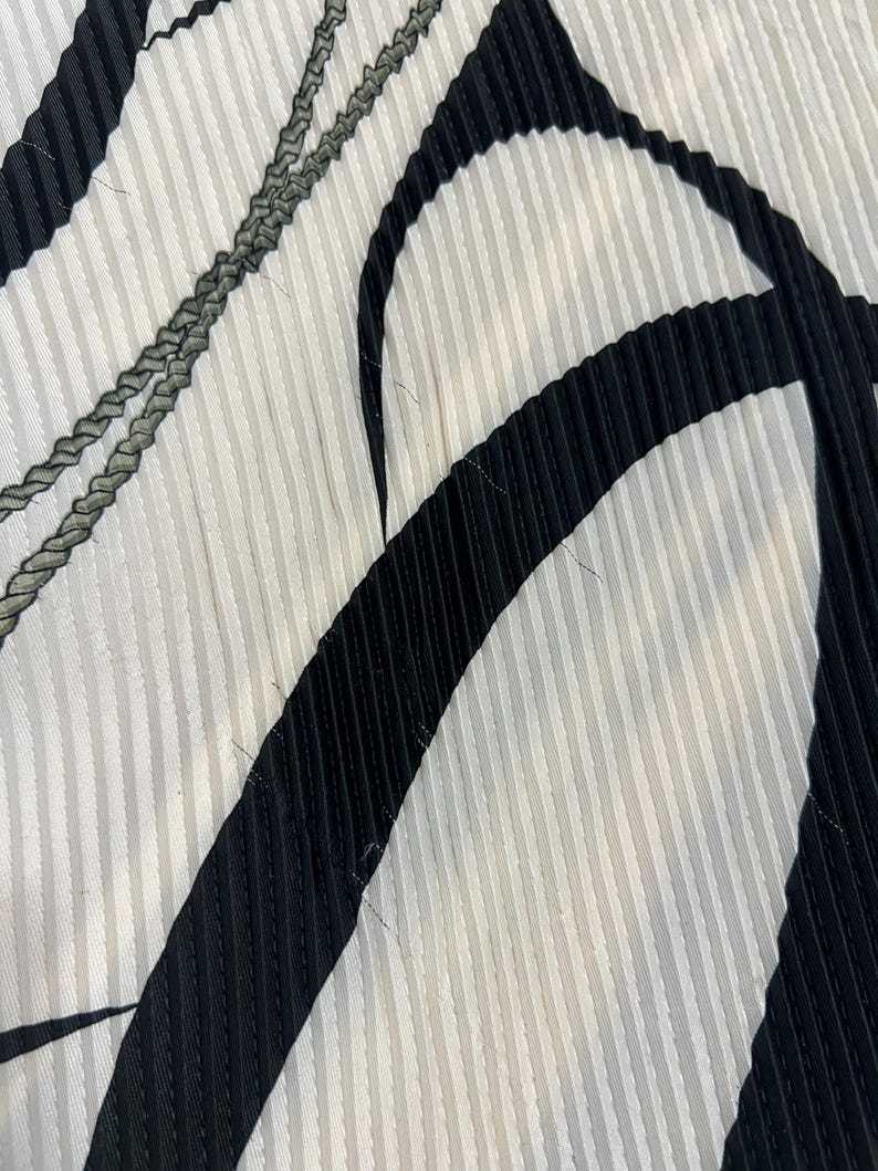 May include: Close-up of a fabric with a pleated design. The fabric features a pattern of vertical white stripes, with bold black curved shapes and a gray braided detail. The texture is clearly visible, highlighting the pleats.