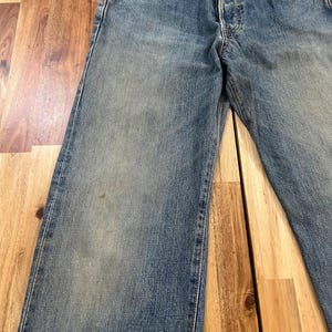 May include: Faded blue denim jeans with a button fly and classic five-pocket design. The jeans show signs of wear, with subtle fading and a relaxed fit. The image showcases the front of the jeans, highlighting the details of the pockets and stitching.