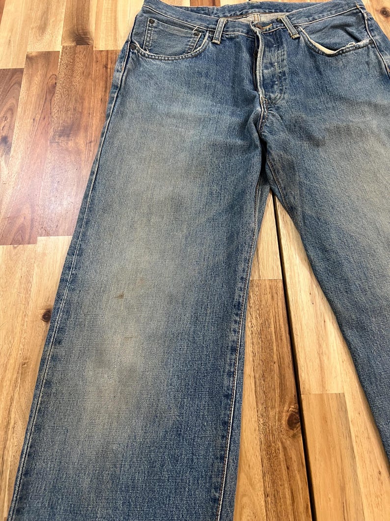 May include: Faded blue denim jeans with a button fly and classic five-pocket design. The jeans show signs of wear, with subtle fading and a relaxed fit. The image showcases the front of the jeans, highlighting the details of the pockets and stitching.