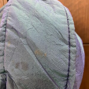 K&ouml;nnte beinhalten: Nahaufnahme eines Abschnitts eines blau-grauen, gesteppten Stoffes. Der Stoff hat einen Glanz und sichtbare N&auml;hte. Auf dem Stoff sind mehrere braune Flecken zu sehen. Der Artikel scheint ein Kleidungsst&uuml;ck oder eine Tasche zu sein.
