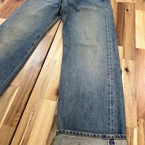 May include: Faded blue denim jeans with a button fly. The jeans have a relaxed fit and are cuffed at the ankles, revealing a red and white selvedge detail. The denim shows signs of wear.
