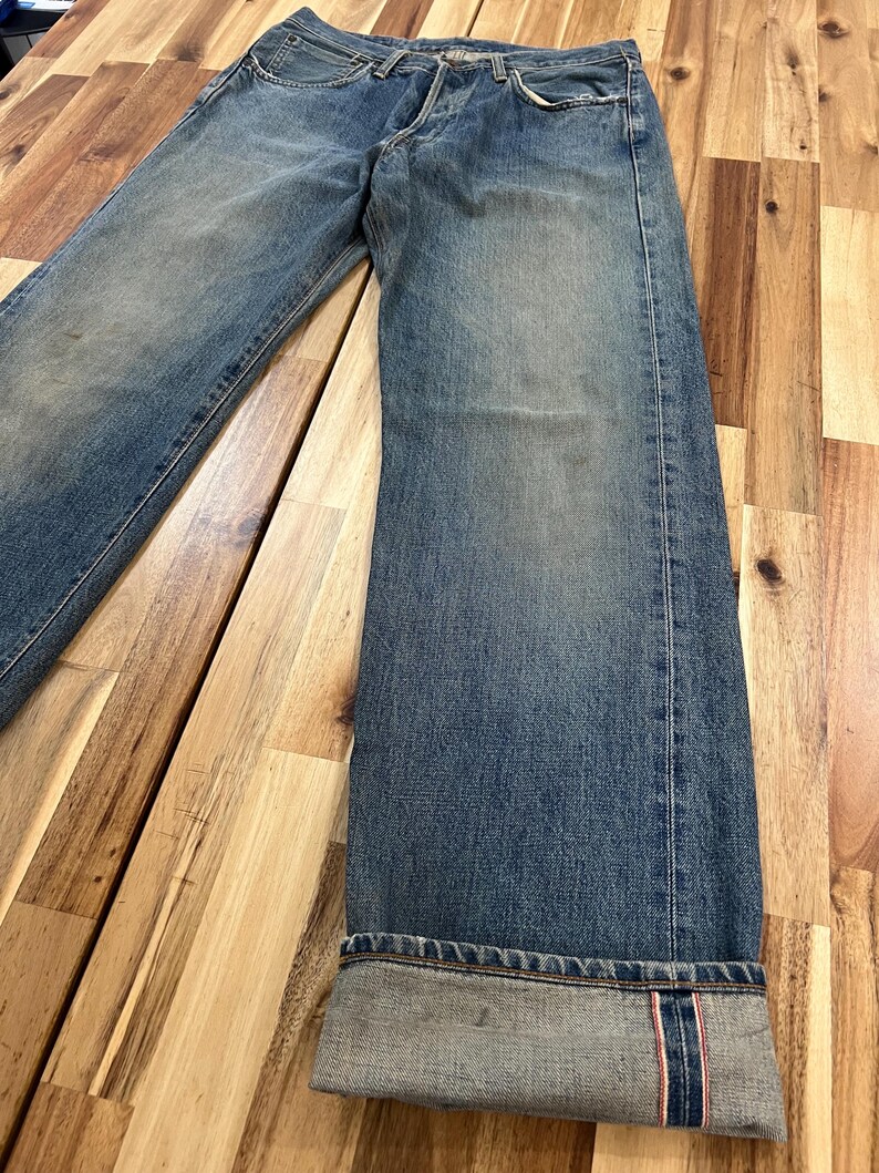 May include: Faded blue denim jeans with a button fly. The jeans have a relaxed fit and are cuffed at the ankles, revealing a red and white selvedge detail. The denim shows signs of wear.