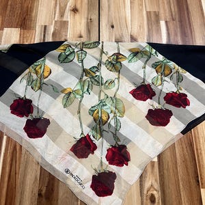 May include: A square scarf featuring a floral design of red roses with green leaves and stems. The scarf has a striped pattern in beige and white, with a black border. The brand name "PAOLOGUCCI" is visible.