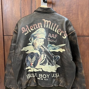 May include: Brown leather bomber jacket with a graphic on the back featuring a musician playing a trombone, the text "Glenn Miller's AAF Band," and "BUGE BOY U.S.A.". The jacket has a green ribbed hem and cuffs.