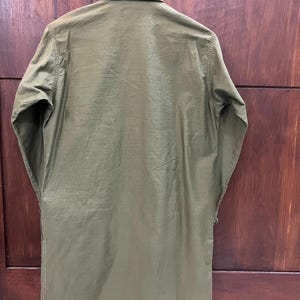 May include: An olive green long-sleeved shirt with a pointed collar and a button-down front. The garment features a straight hemline and a smooth, solid-colored fabric construction, presented from a rear-facing perspective while hanging on a hanger.
