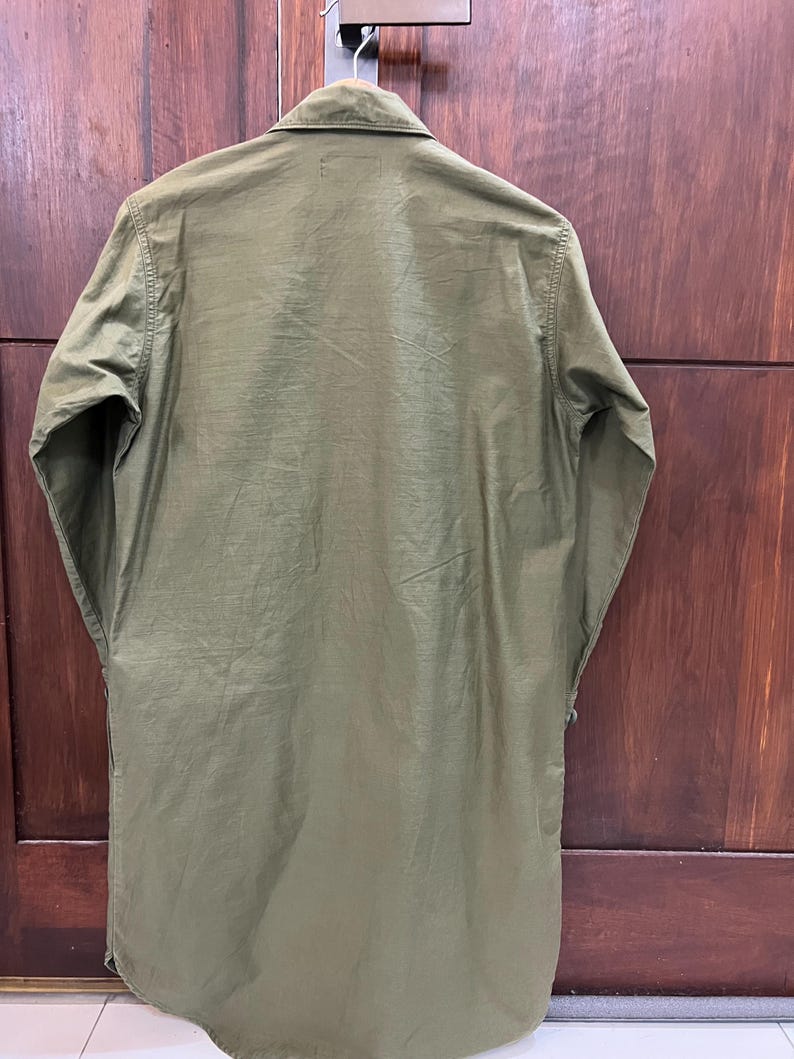 May include: An olive green long-sleeved shirt with a pointed collar and a button-down front. The garment features a straight hemline and a smooth, solid-colored fabric construction, presented from a rear-facing perspective while hanging on a hanger.