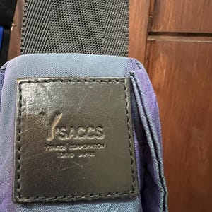 K&ouml;nnte beinhalten: Nahaufnahme einer blau-violett gemusterten Tasche mit schwarzem Riemen. Ein schwarzes Lederetikett ist auf die Tasche gen&auml;ht und zeigt den Markennamen "YSACCS" und "YSACCS CORPORATION TOKYO JAPAN". Der Riemen der Tasche ist schwarz.