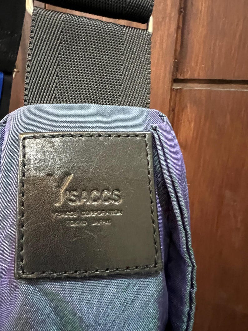 K&ouml;nnte beinhalten: Nahaufnahme einer blau-violett gemusterten Tasche mit schwarzem Riemen. Ein schwarzes Lederetikett ist auf die Tasche gen&auml;ht und zeigt den Markennamen "YSACCS" und "YSACCS CORPORATION TOKYO JAPAN". Der Riemen der Tasche ist schwarz.