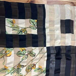 May include: A rectangular scarf with a floral rose pattern. The scarf features a patchwork design with alternating stripes of black, brown, and beige. The floral pattern includes red roses and green leaves.