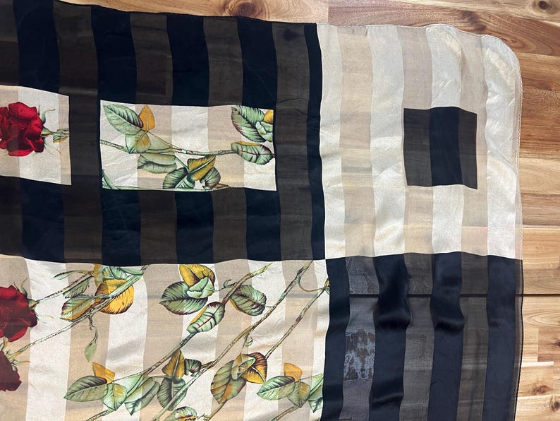 May include: A rectangular scarf with a floral rose pattern. The scarf features a patchwork design with alternating stripes of black, brown, and beige. The floral pattern includes red roses and green leaves.