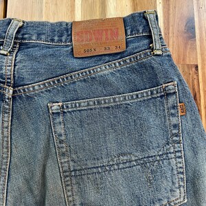 May include: Close-up of blue denim jeans with a back pocket and a brown leather patch. The patch reads "EDWIN" and includes the numbers "505 X 33 34". The jeans have a faded, worn look.