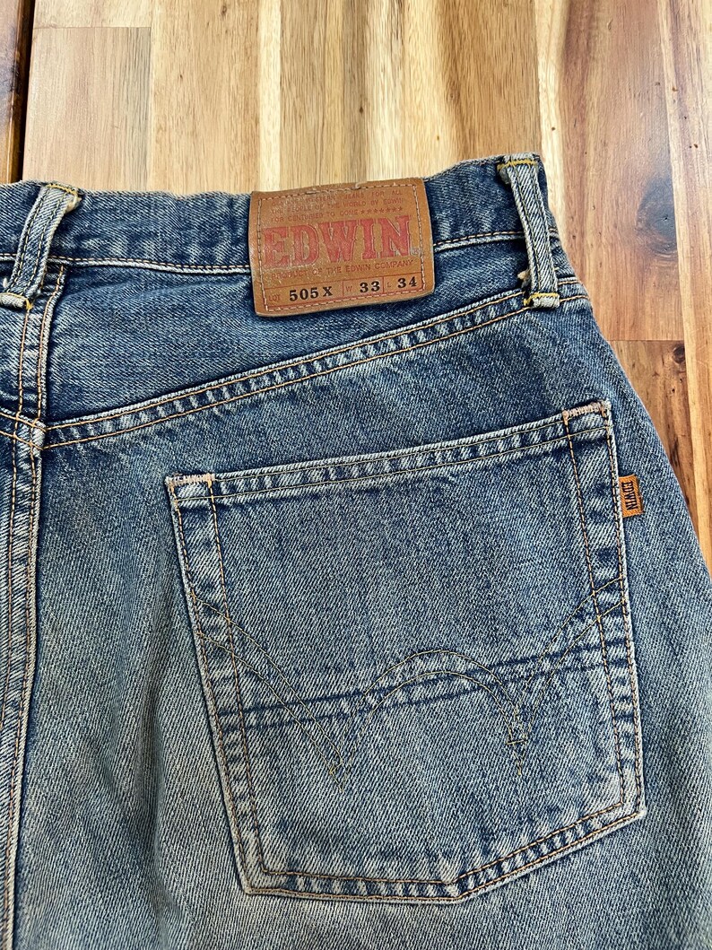 May include: Close-up of blue denim jeans with a back pocket and a brown leather patch. The patch reads "EDWIN" and includes the numbers "505 X 33 34". The jeans have a faded, worn look.
