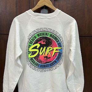 May include: White crew neck sweatshirt with a vibrant surf design. The back features the word "SURF" in yellow and pink, a surfer silhouette, and the words "Waikiki" and "Hawaii North Shore" in a circular pattern. A vintage style.