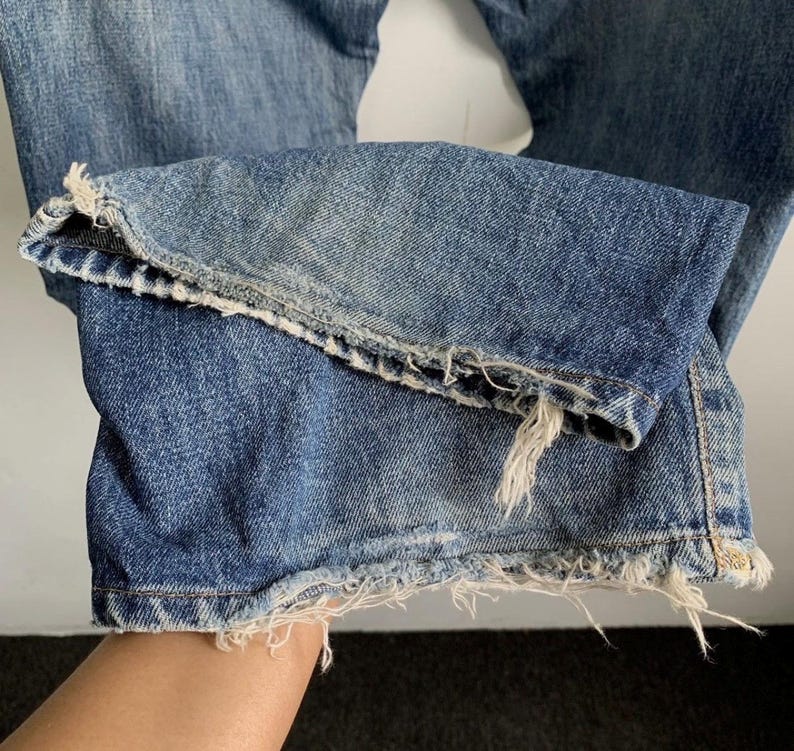 Peut inclure: Gros plan sur l'ourlet d'un jean en denim bleu. Le jean pr&eacute;sente un bord effiloch&eacute; et vieilli, avec des fils blancs visibles. Le denim montre des signes d'usure et de d&eacute;coloration, lui donnant un aspect vintage.
