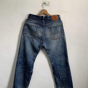 Peut inclure: Un jean en denim bleu vintage avec un aspect us&eacute;, avec deux poches arri&egrave;re et un patch en cuir. Le jean est suspendu &agrave; un cintre en bois contre un mur blanc. Le denim a un aspect d&eacute;lav&eacute; et vieilli.