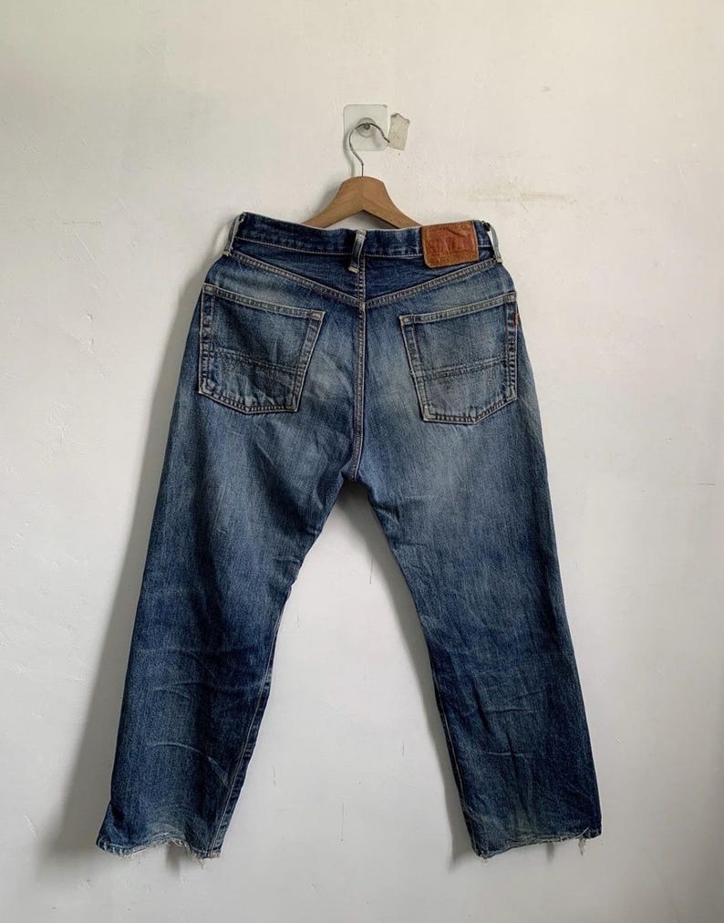 Peut inclure: Un jean en denim bleu vintage avec un aspect us&eacute;, avec deux poches arri&egrave;re et un patch en cuir. Le jean est suspendu &agrave; un cintre en bois contre un mur blanc. Le denim a un aspect d&eacute;lav&eacute; et vieilli.