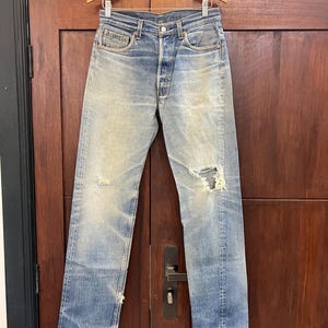 May include: Light blue denim jeans featuring a faded wash, a button fly closure, and distressed detailing including a prominent tear on the left knee and frayed hems. The jeans have a classic five-pocket design with copper-toned rivets.