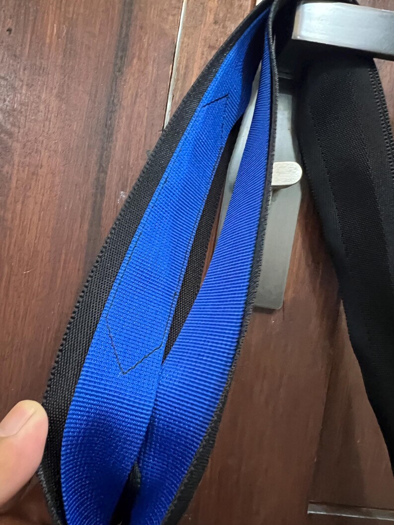 K&ouml;nnte beinhalten: Nahaufnahme eines schwarz-blauen Gurtes mit silbernem Metallverschluss. Der Gurt hat eine blaue Mitte und schwarze R&auml;nder. Der Gurt besteht aus einem gewebten Material.
