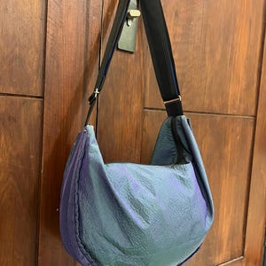 K&ouml;nnte beinhalten: Eine gro&szlig;e, schillernde Umh&auml;ngetasche mit schwarzem Riemen. Der Taschenkorpus hat ein strukturiertes, zerknittertes Aussehen, wobei die Farben zwischen Blau, Gr&uuml;n und Lila wechseln. Der Riemen ist schwarz mit blauen Akzenten. Die Tasche h&auml;ngt an einer Holzt&uuml;r.