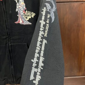 Pu&ograve; includere: Felpa con cappuccio nera con zip e grafica di ispirazione giapponese. Presenta un design di geisha e la scritta "Definite Stance" sul petto. La manica mostra una fenice e una scritta.