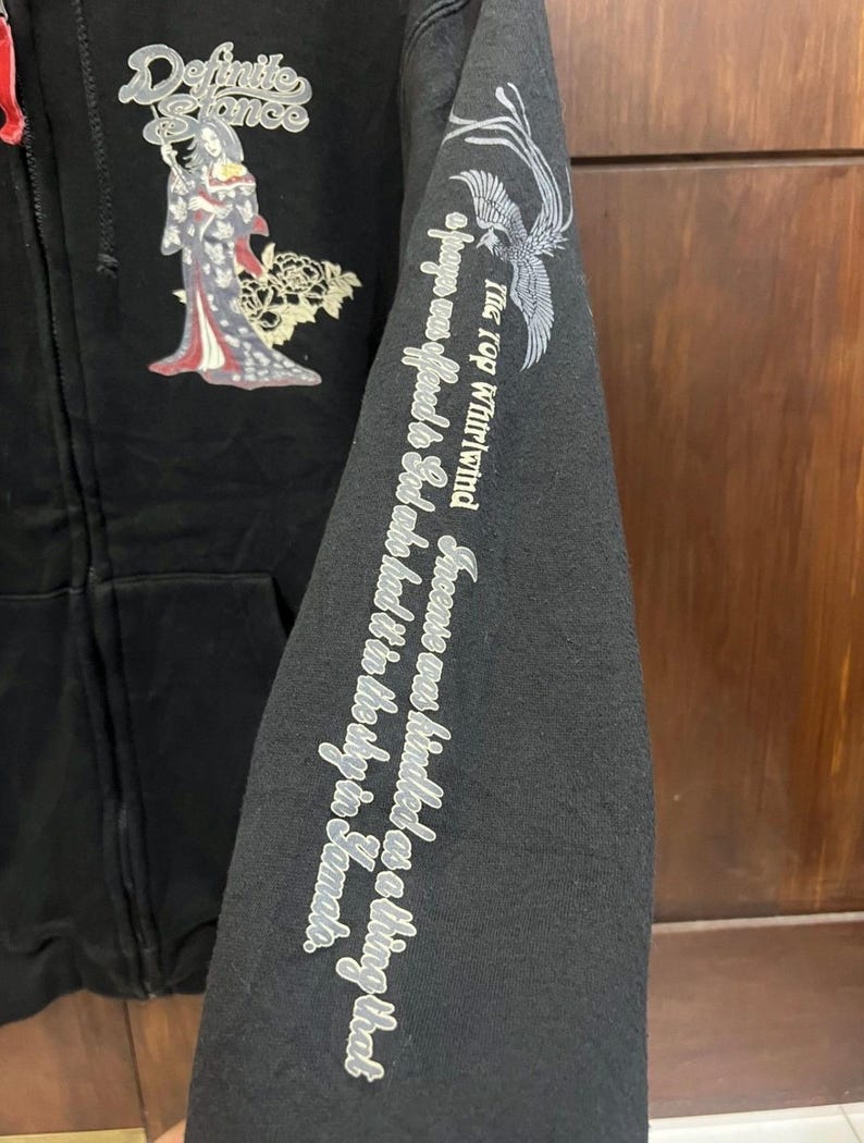 Pu&ograve; includere: Felpa con cappuccio nera con zip e grafica di ispirazione giapponese. Presenta un design di geisha e la scritta "Definite Stance" sul petto. La manica mostra una fenice e una scritta.