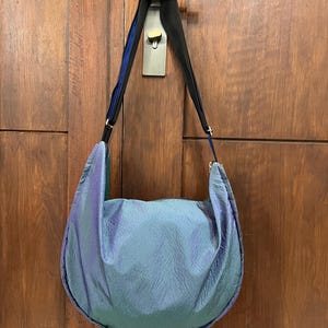 K&ouml;nnte beinhalten: Eine gro&szlig;e, schillernde Umh&auml;ngetasche in Blau- und Gr&uuml;nt&ouml;nen h&auml;ngt an einer Holzt&uuml;r. Die Tasche hat eine halbmondf&ouml;rmige Form und einen schwarzen Riemen mit blauen Akzenten. Das Material der Tasche hat ein strukturiertes, zerknittertes Aussehen.