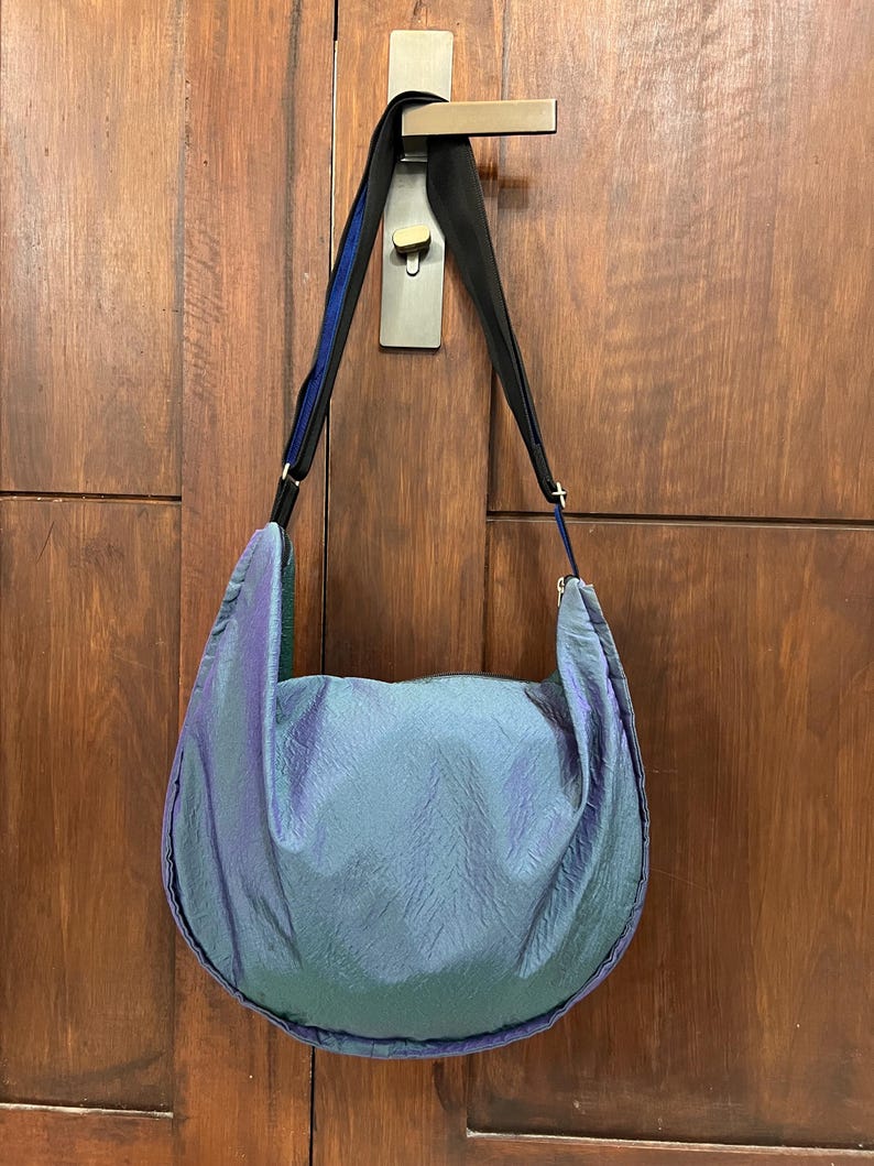 K&ouml;nnte beinhalten: Eine gro&szlig;e, schillernde Umh&auml;ngetasche in Blau- und Gr&uuml;nt&ouml;nen h&auml;ngt an einer Holzt&uuml;r. Die Tasche hat eine halbmondf&ouml;rmige Form und einen schwarzen Riemen mit blauen Akzenten. Das Material der Tasche hat ein strukturiertes, zerknittertes Aussehen.