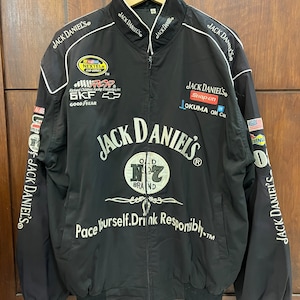 Puede incluir: Chaqueta de carreras negra con ribetes blancos y el logotipo de Jack Daniel's. La chaqueta tiene varios logotipos de patrocinadores, incluidos NASCAR, SKF, Snap-on y Okuma. El texto "Pace Yourself. Drink Responsibly" está impreso en la parte delantera de la chaqueta.
