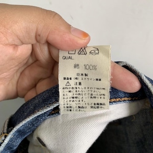 Peut inclure: Une &eacute;tiquette blanche avec du texte et des symboles japonais, tenue par une personne. L'&eacute;tiquette indique "QUAL" en haut, suivi de "綿 100%" et de texte japonais. L'&eacute;tiquette comprend &eacute;galement des instructions d'entretien et un avertissement concernant le transfert de couleur.