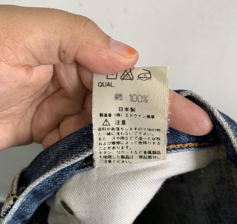 Peut inclure: Une &eacute;tiquette blanche avec du texte et des symboles japonais, tenue par une personne. L'&eacute;tiquette indique "QUAL" en haut, suivi de "綿 100%" et de texte japonais. L'&eacute;tiquette comprend &eacute;galement des instructions d'entretien et un avertissement concernant le transfert de couleur.