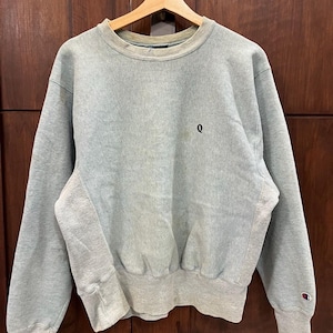 May include: A pale grey sweatshirt with a crew neck and long sleeves. The sweatshirt features a small black "Q" embroidered on the chest and a Champion logo on the left cuff. The fabric looks to be a soft, comfortable material.