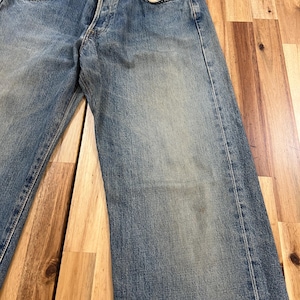 May include: Faded blue denim jeans with a button fly and classic five-pocket design. The jeans show signs of wear, with slight fading and distressing, giving them a vintage look. The jeans are laid flat on a wooden surface.