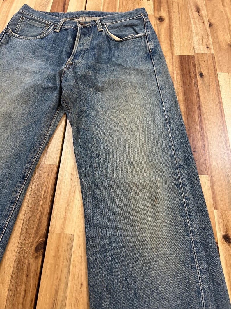 May include: Faded blue denim jeans with a button fly and classic five-pocket design. The jeans show signs of wear, with slight fading and distressing, giving them a vintage look. The jeans are laid flat on a wooden surface.