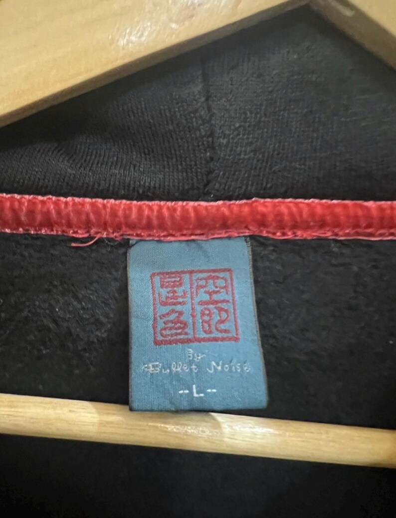 Pu&ograve; includere: Primo piano di una felpa con cappuccio nera con finiture rosse sul cappuccio. Un'etichetta blu con caratteri cinesi rossi e la scritta "by Bullet Noise" &egrave; visibile. L'etichetta indica anche una taglia "-L-".
