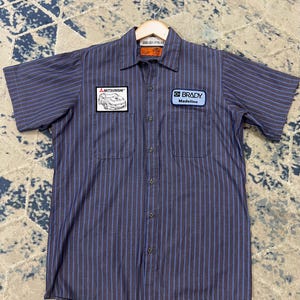 May include: A short-sleeved, button-down work shirt in a blue and purple striped pattern. The shirt has two chest pockets and patches that read "MITSUBISHI" and "BRADY Madeline". The shirt is hanging on a wooden hanger.