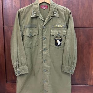 May include: An olive green long-sleeve button-down shirt featuring a U.S. ARMY patch, an AIRBORNE eagle patch, and star collar insignias. The shirt has two chest pockets with button flaps and a red interior neck label with white text.