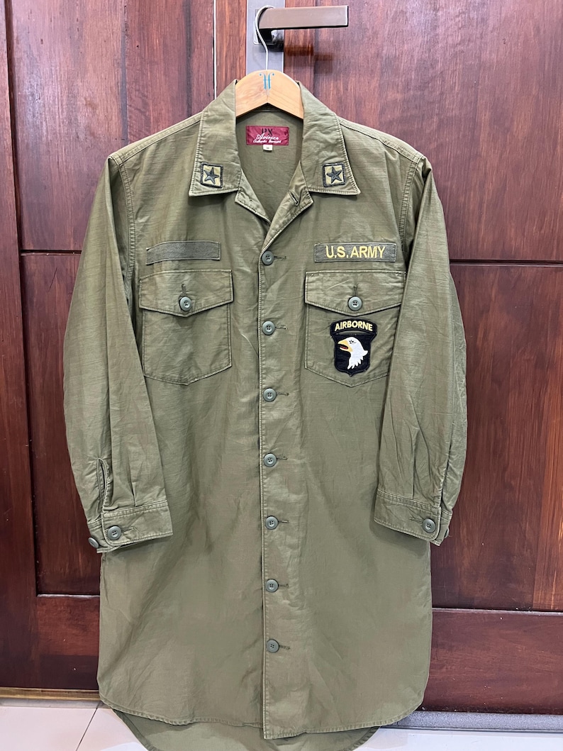 May include: An olive green long-sleeve button-down shirt featuring a U.S. ARMY patch, an AIRBORNE eagle patch, and star collar insignias. The shirt has two chest pockets with button flaps and a red interior neck label with white text.