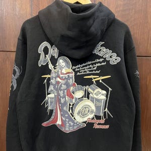May include: Black hoodie with a graphic on the back. The design features a woman in a kimono playing drums, with the words "Reverence" and "Fundamental Reverence". The sleeves have decorative elements. The hoodie is hanging on a wooden hanger.