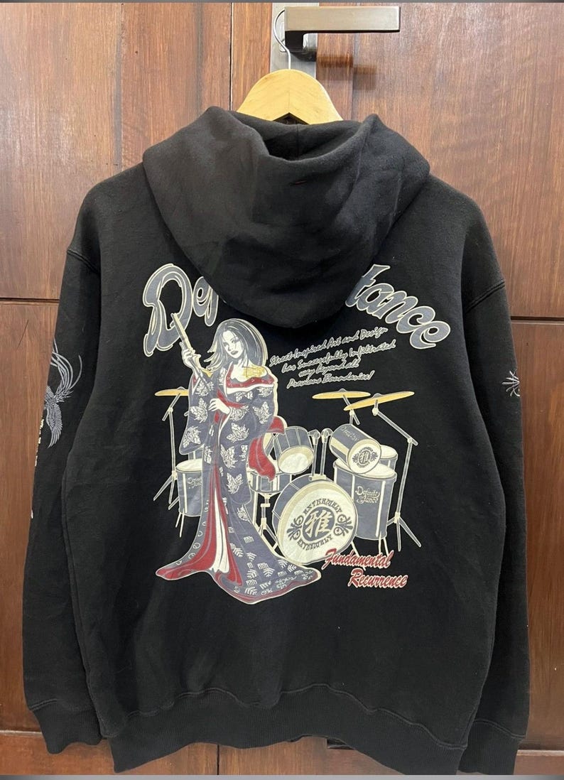 Pu&ograve; includere: Felpa con cappuccio nera con grafica sul retro. Il design presenta una donna in kimono che suona la batteria, con le parole "Reverence" e "Fundamental Reverence". Le maniche hanno elementi decorativi.