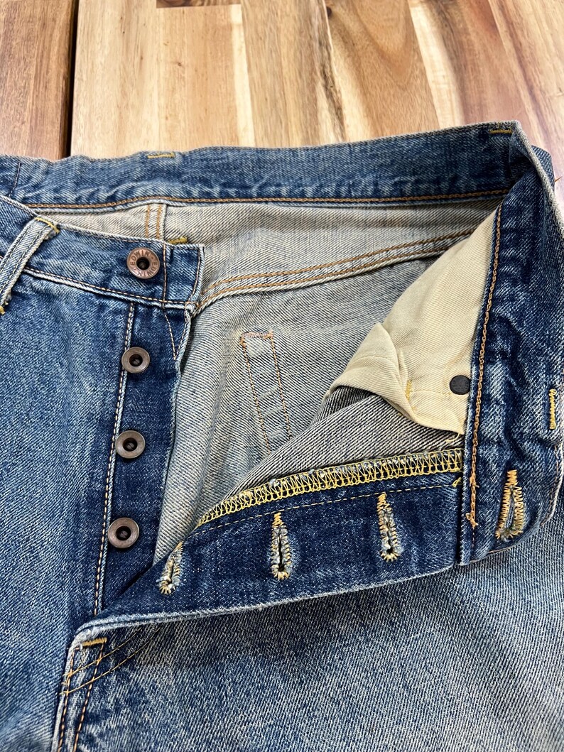 May include: Close-up of blue denim jeans with a button fly. The jeans show signs of wear and fading, with visible stitching in yellow thread. The buttons are a dark, metallic color.